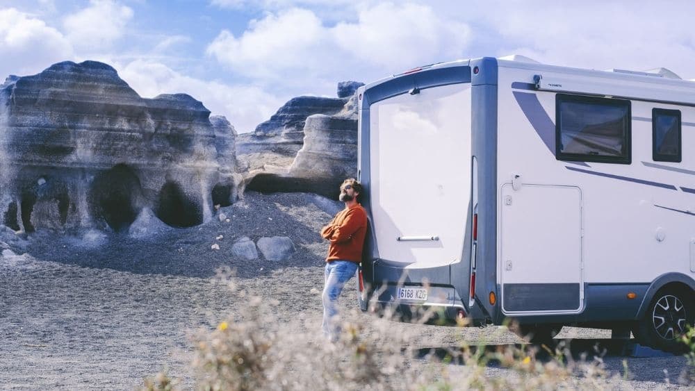 Person leaning against a parked RV in an open field.