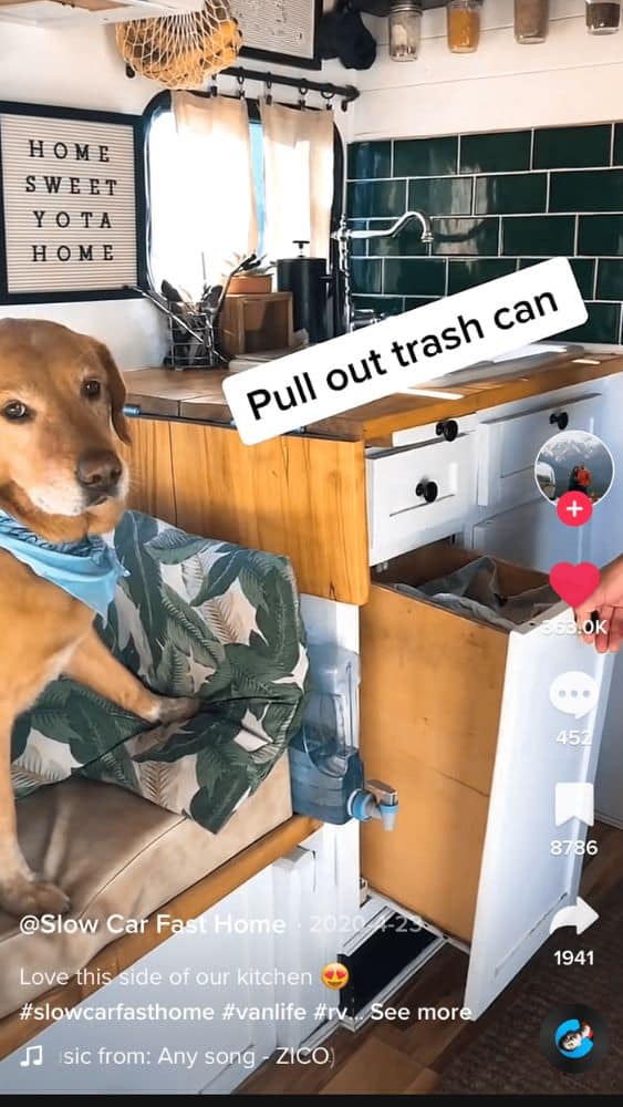 Screenshot from @Slow Car Fast Home's TikTok video that shows a pullout trash can under a kitchen counter. A gold dog with a blue bandana sits on a bench on the left side of the image.