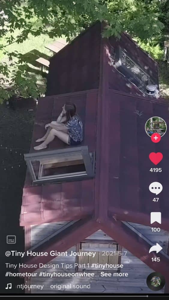 Screenshot of @Tiny House Giant Journey's TikTok video showing the roof of a tiny house from above. A person sits next to an open skylight.
