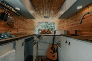 Wood panel interior of a customized van with wood countertops, sink, fridge, stove, and a custom-made bed by the window