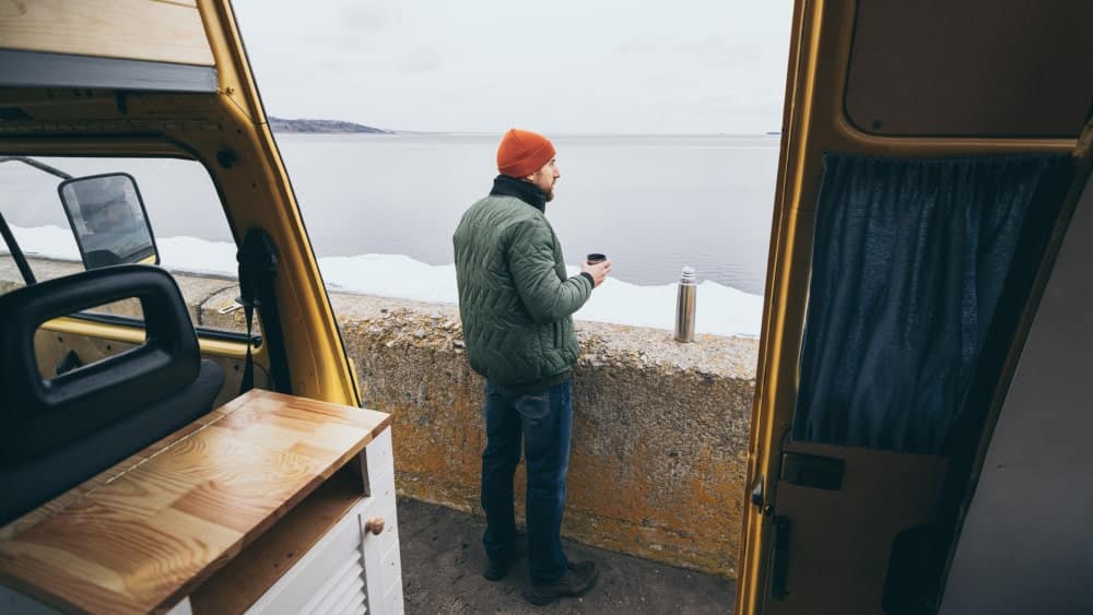 Young man in red cap stands next to van door drinking tea in front of lake in winter (snow on the ground)